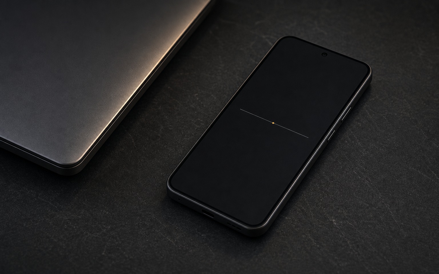 HushPhone beside a closed laptop on dark graphite, its screen nearly empty with a single amber status dot — a quiet channel, open and listening.